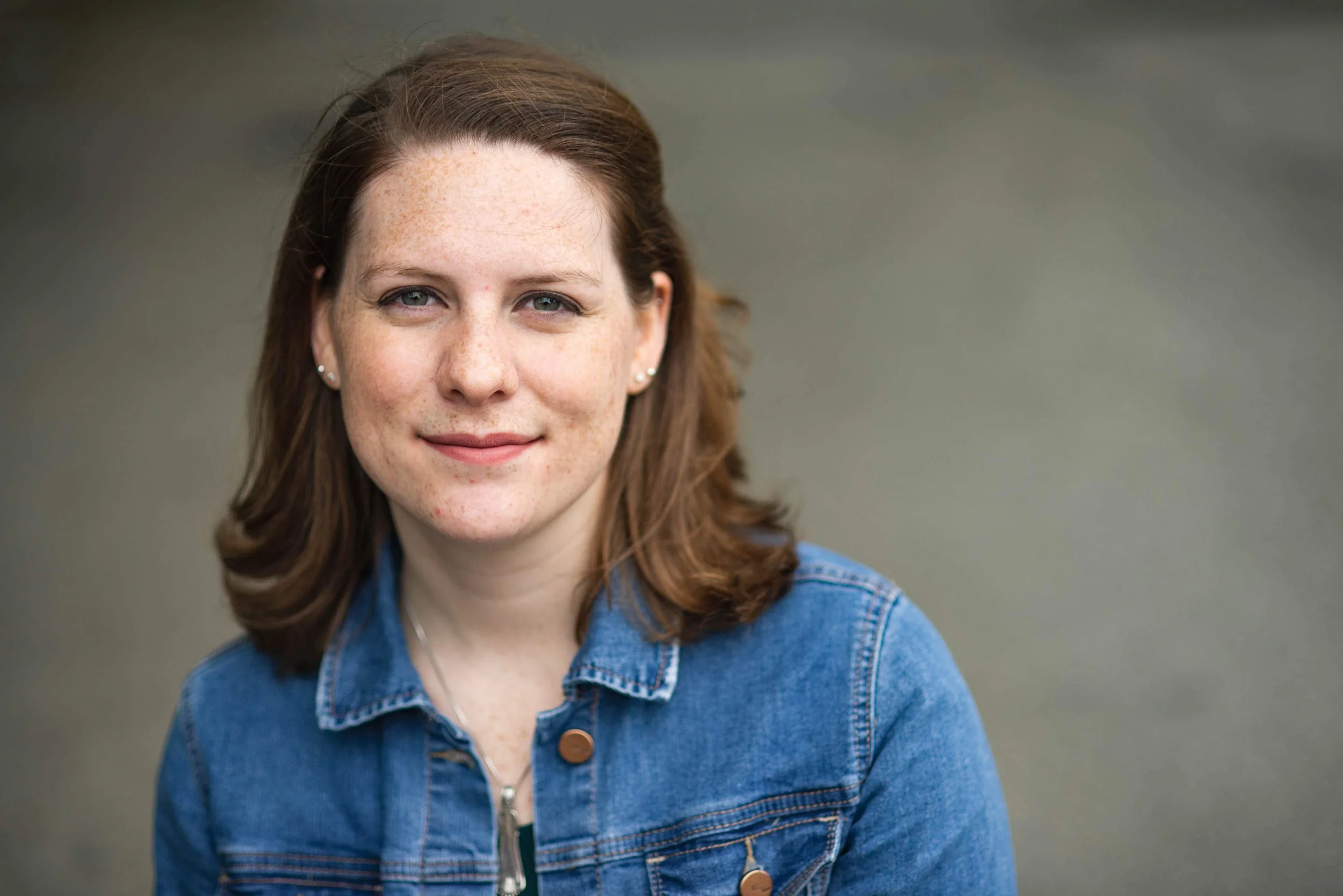 Portrait of the author with auburn hair, green eyes, and fair skin with freckles, wearing a denim jacket and small earrings, standing outdoors with a blurred green background.