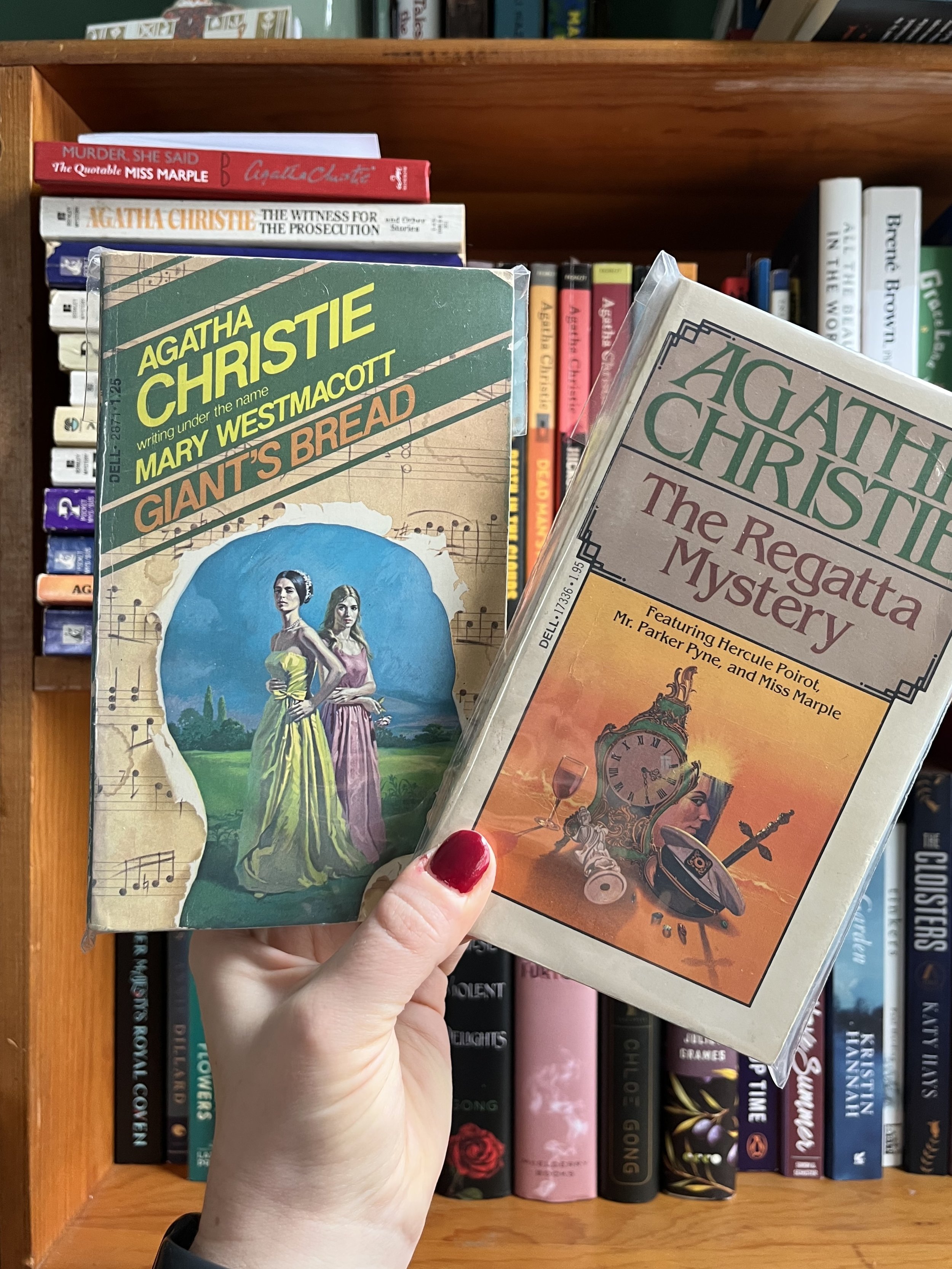 Hand holding two Agatha Christie books, 'Giant's Bread' and 'The Regatta Mystery,' in front of a bookshelf with other various books.