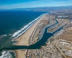 Aerial view of a coastal harbor with a breakwater, marina, and shoreline, extending along the beachfront with waters and land on either side.