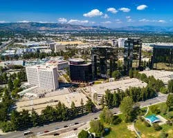 Aerial view of a cityscape with tall modern buildings, green spaces, and a pool, under a blue sky with scattered clouds.