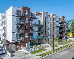 Modern multi-story apartment building with balconies, windows, and a sidewalk in front, under a clear blue sky.