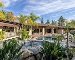 A backyard with a swimming pool, surrounded by rocks and lush tropical plants, with a house in the background and a partly cloudy sky overhead.