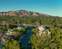 A neighborhood with houses surrounded by trees, a main road running through, and a mountain in the background under a blue sky.