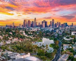 Skyline of a city at sunset with tall skyscrapers, a river, and green parks in the foreground.