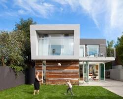 Modern two-story house with large windows and a balcony, blue sky, trees, and a lawn with three people playing with a dog.