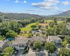 Residential neighborhood with houses and trees, and a large green park or golf course in the background under a partly cloudy sky.