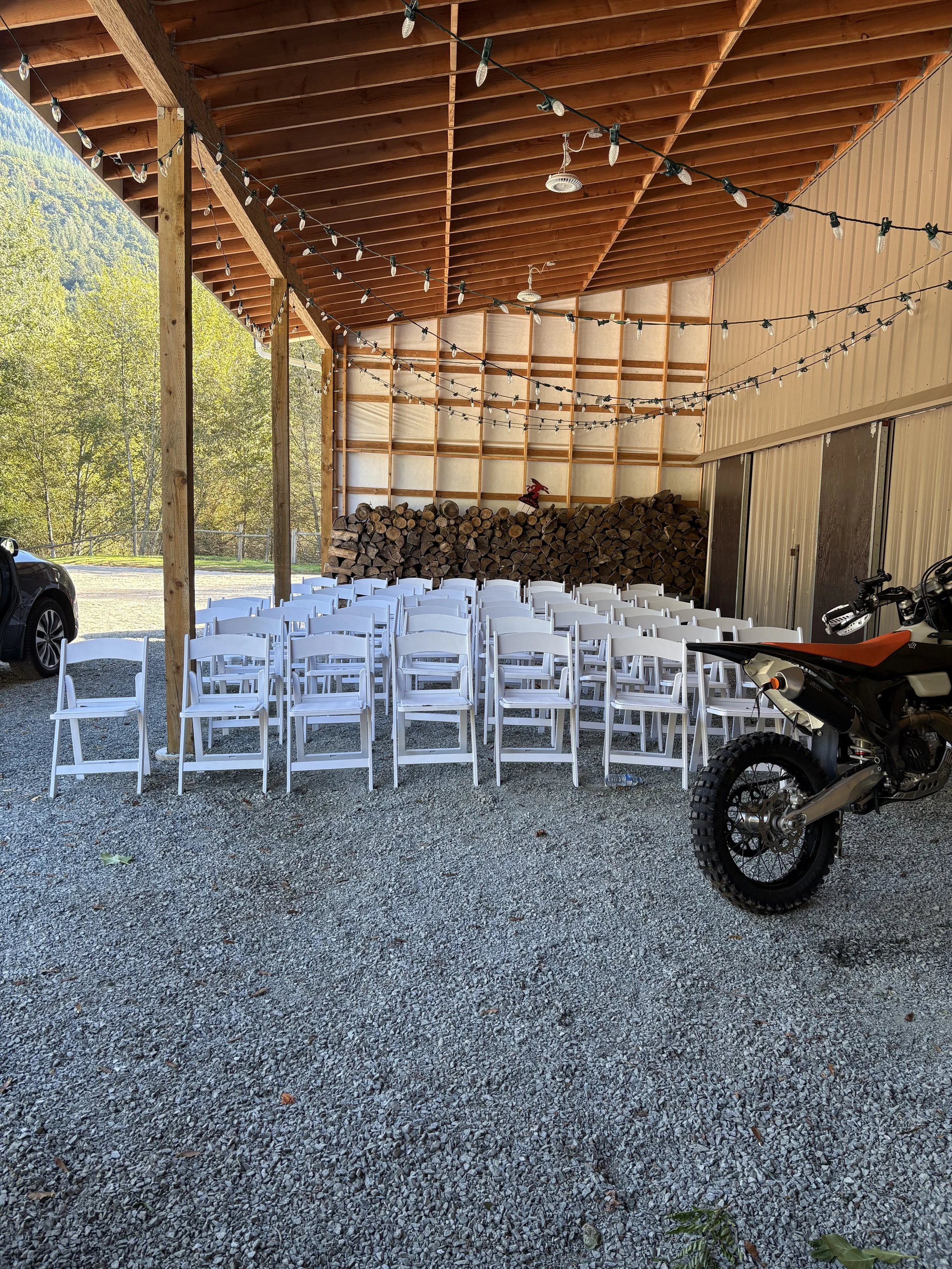 Outdoor covered patio with white chairs arranged in rows, string lights hanging from wooden beams, logs stacked against the back wall, a motorcycle parked to the right, and trees visible in the background.