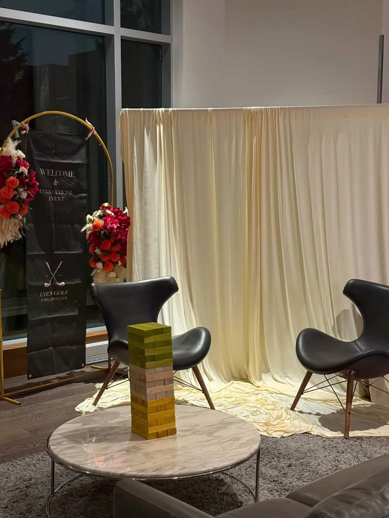 Interior with a cream-colored curtain backdrop, two black modern chairs, a round marble table with a Jenga game stacked on top, and floral arrangements with red and pink flowers near a welcome sign for Eve's official event at Eve's Golf and Wellness Club.