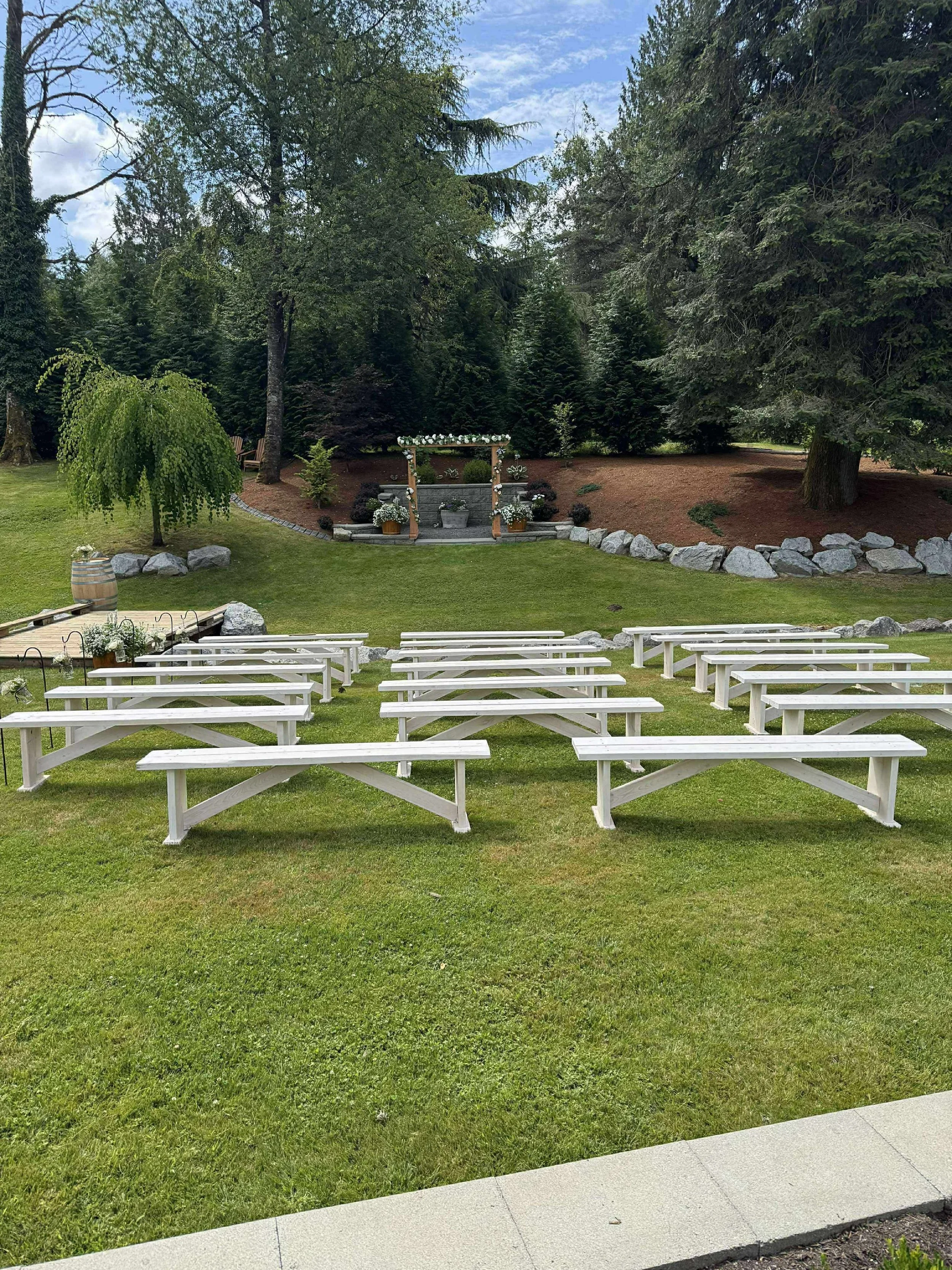 Outdoor wedding setup with white benches facing an altar with floral decorations, surrounded by trees and rocks, on a green lawn under a blue sky.