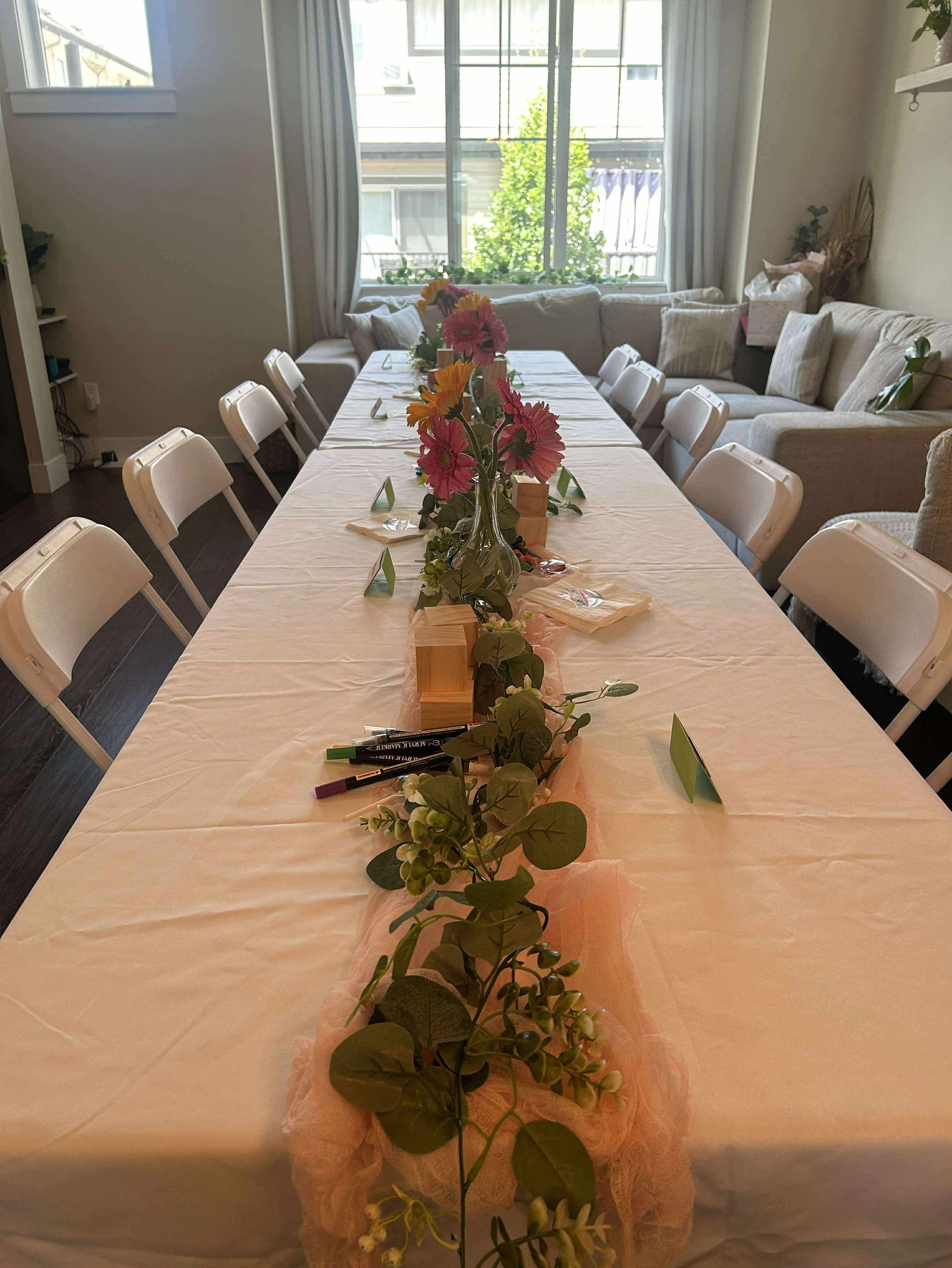 Decorated dining table with a floral centerpiece, set with name cards and pens, in a bright room with large windows and cushioned seating.