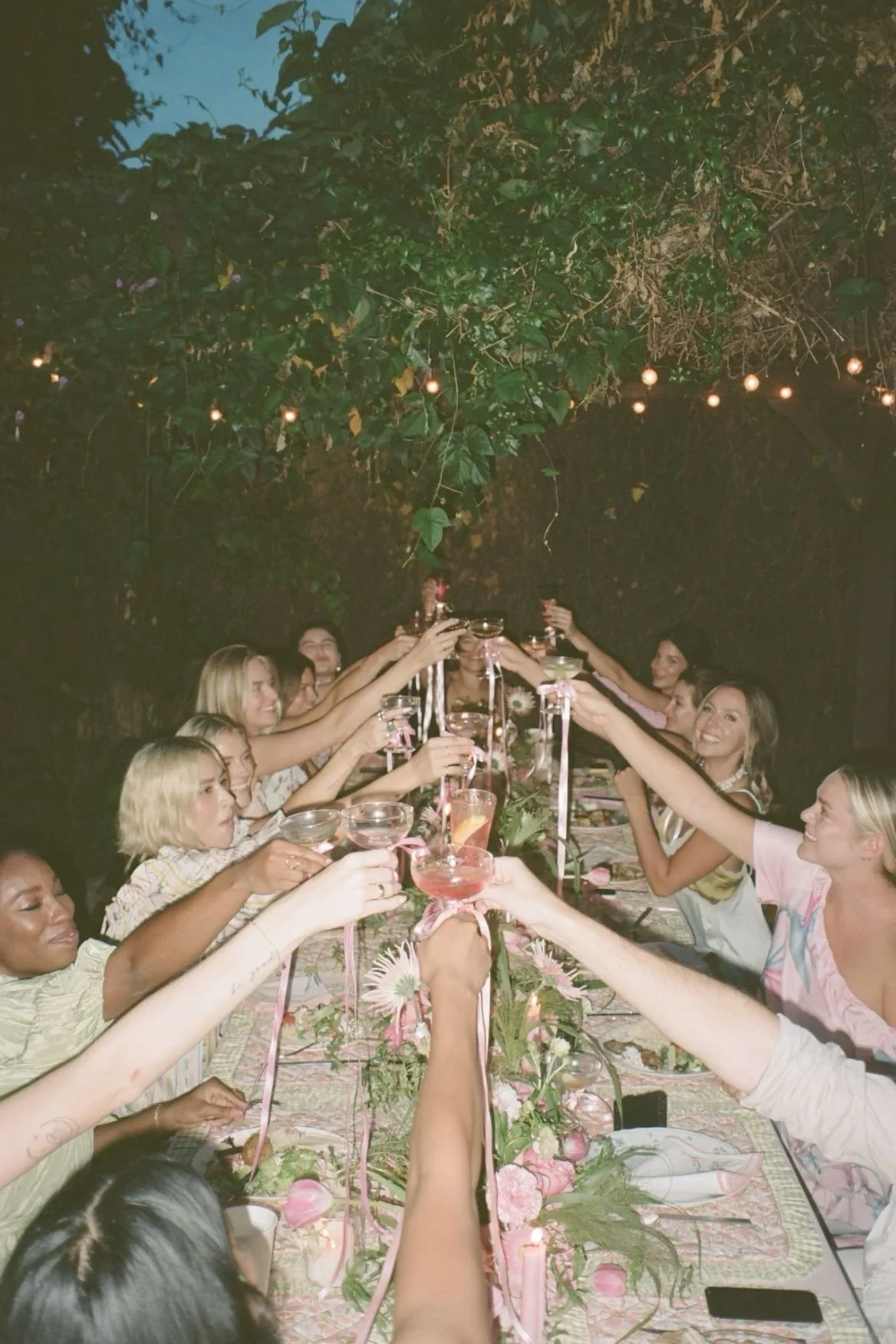 A group of women celebrating at a long outdoor table with floral decorations, raising their drinks in a toast under string lights on a cloudy evening.