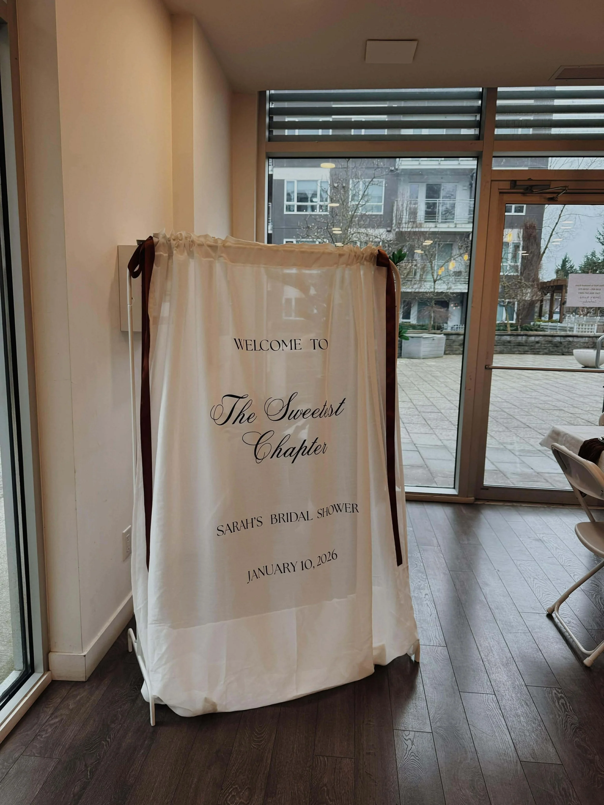 A white fabric sign with black text announcing Sarah's bridal shower on January 10, 2026, at a venue called The Sweetest Chapter, placed inside a room with large windows showing an outdoor patio and modern residential buildings.