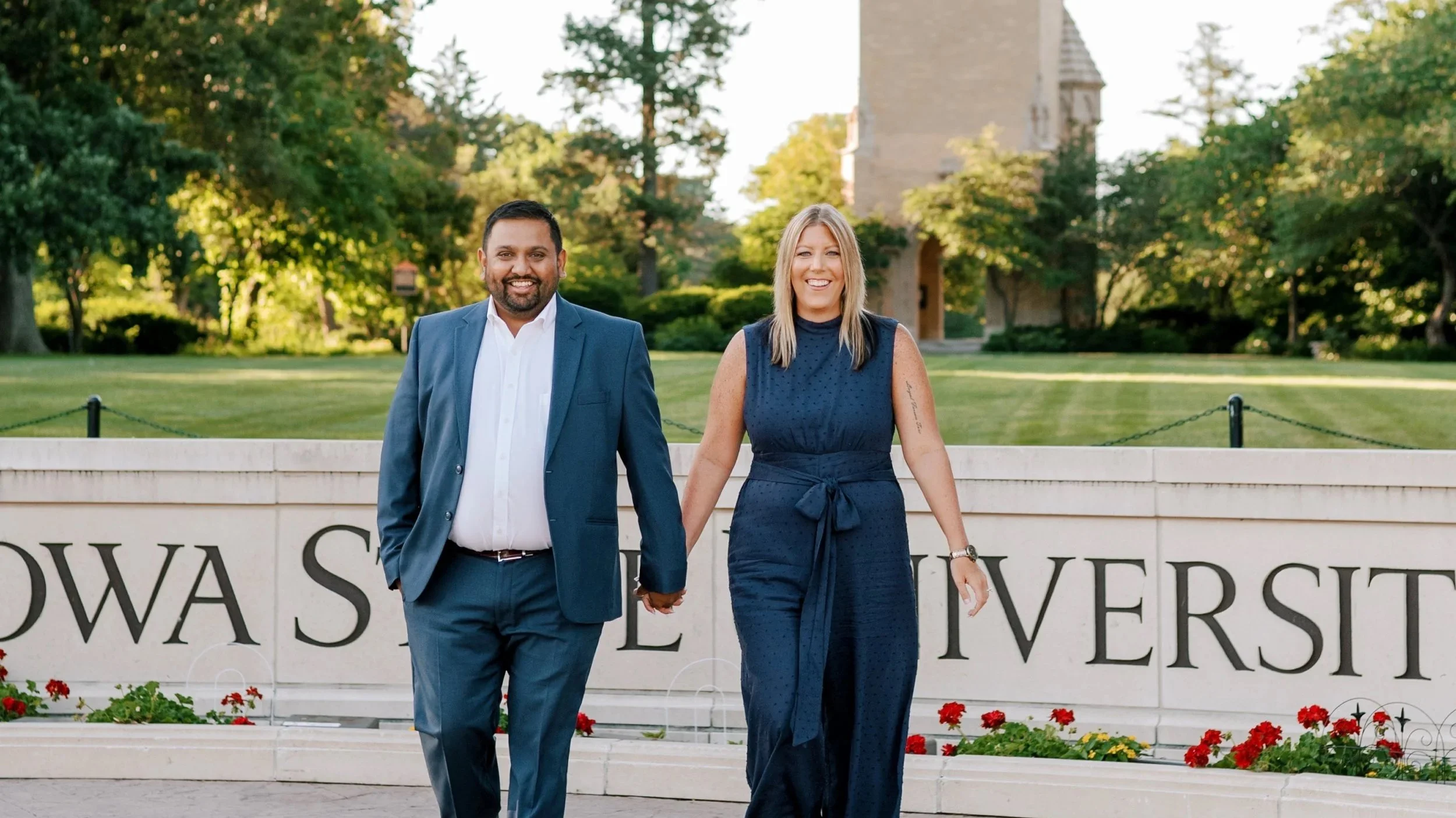 A smiling man and woman holding hands walk in front of a sign that reads 'IOWA STATE UNIVERSITY.' They are outdoors with green trees and a historic building in the background on a sunny day.