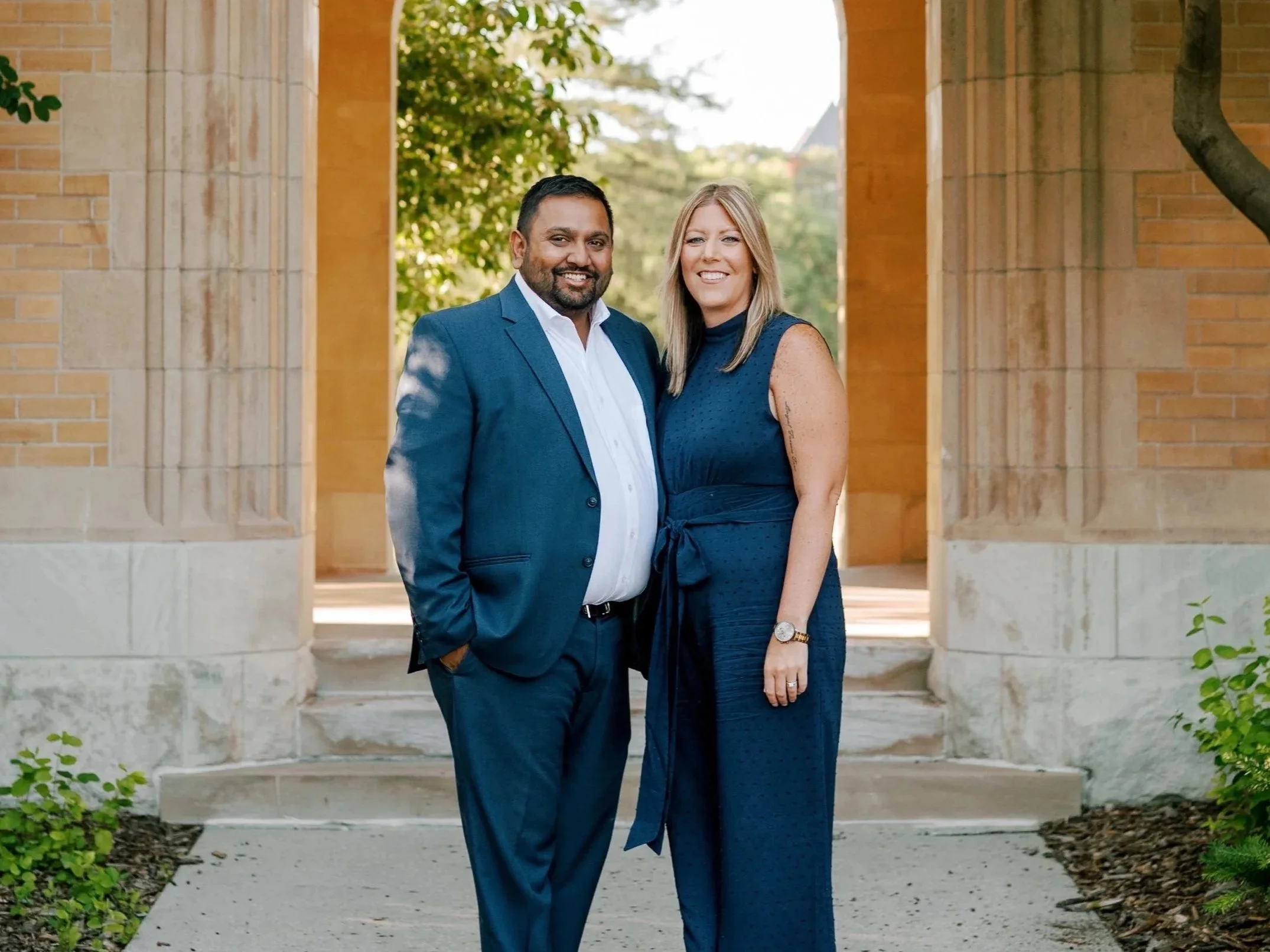 A man and woman standing together outdoors, both smiling, in front of an arched brick structure with trees and greenery in the background.
