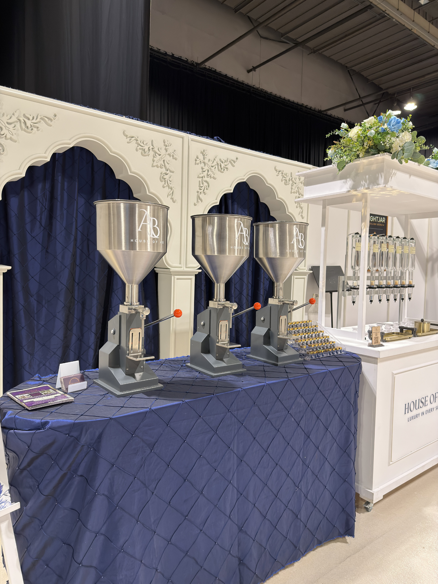 Three large stainless steel dispensers with orange handles on a blue-diamonds quilted tablecloth, with a white display stand with multiple smaller dispensers and decorative flowers on top.