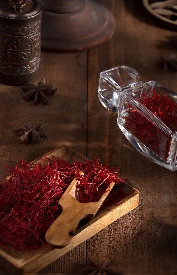 A wooden table holding a small wooden bowl filled with red saffron threads, a glass container with more saffron inside, and star anise spice scattered around.