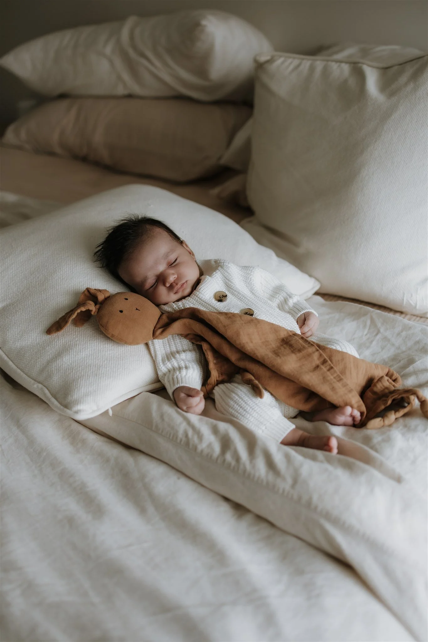 A sleeping baby lying on a cream-colored bed with beige pillows, holding a soft brown stuffed toy.