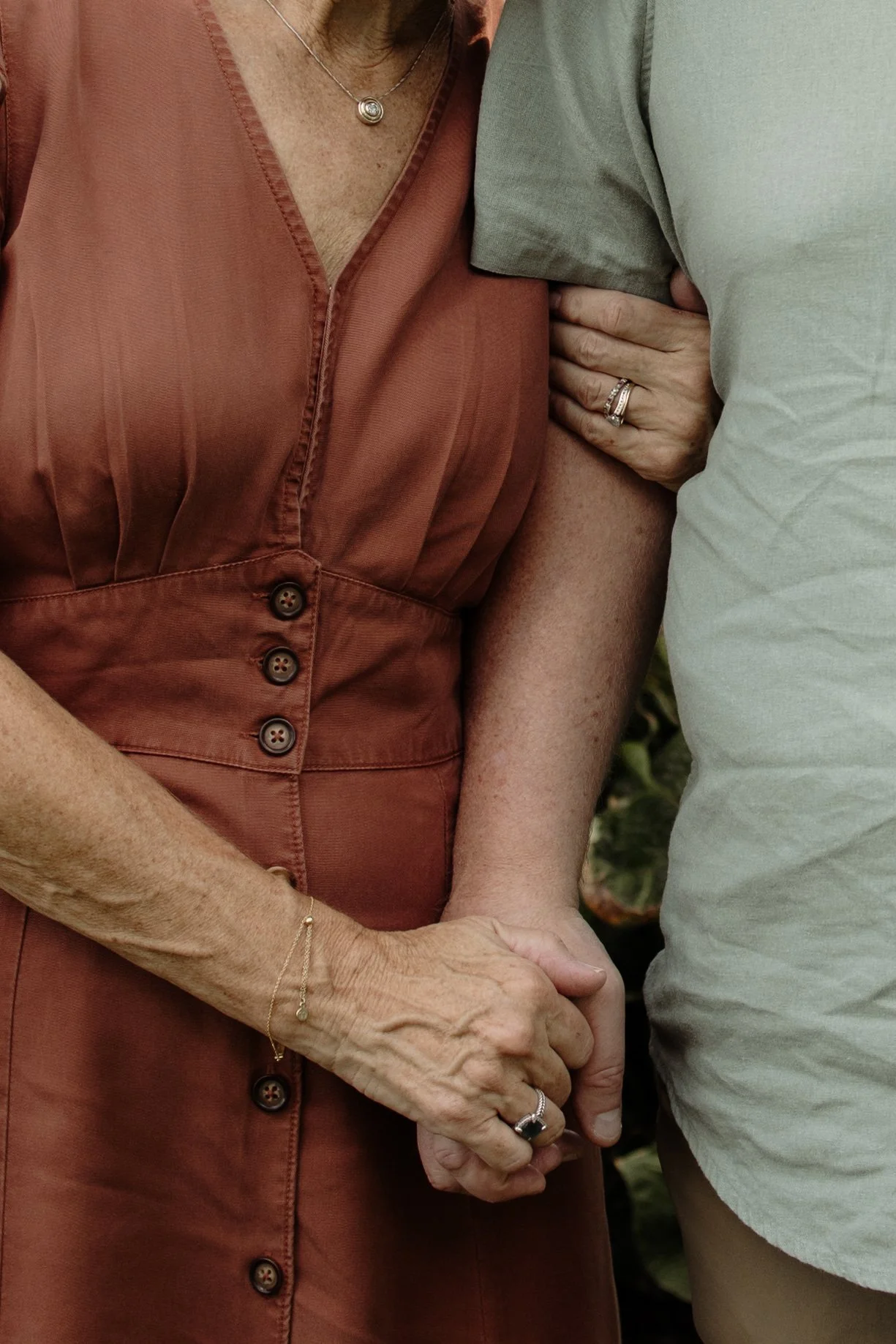 A close-up of two older women holding hands, one wearing a rust-colored dress and the other in a light gray shirt, with the focus on their intertwined hands.