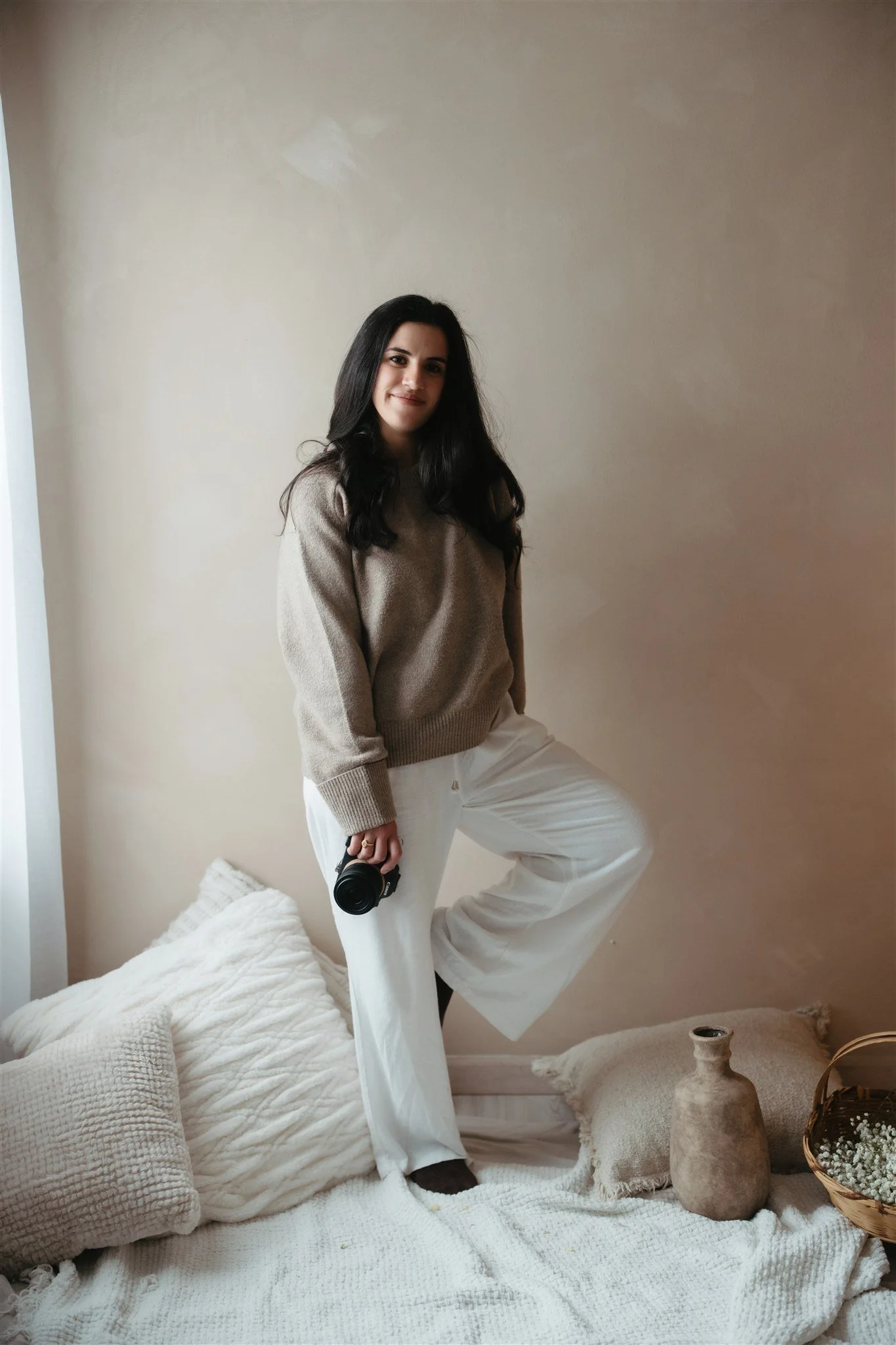 A woman with long dark hair wearing a beige sweater and white pants stands in a cozy, minimalist room, holding a camera, next to a bed with white and beige pillows and rustic decor items.