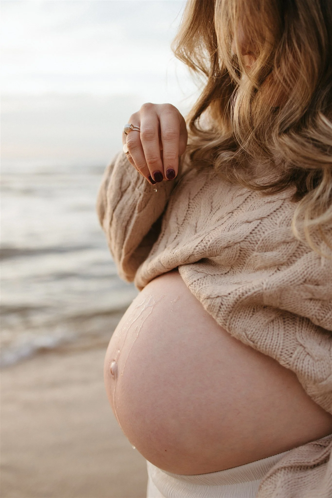 Close-up of a pregnant woman on the beach, wearing a beige sweater, with her hand above her belly, and ocean in the background.