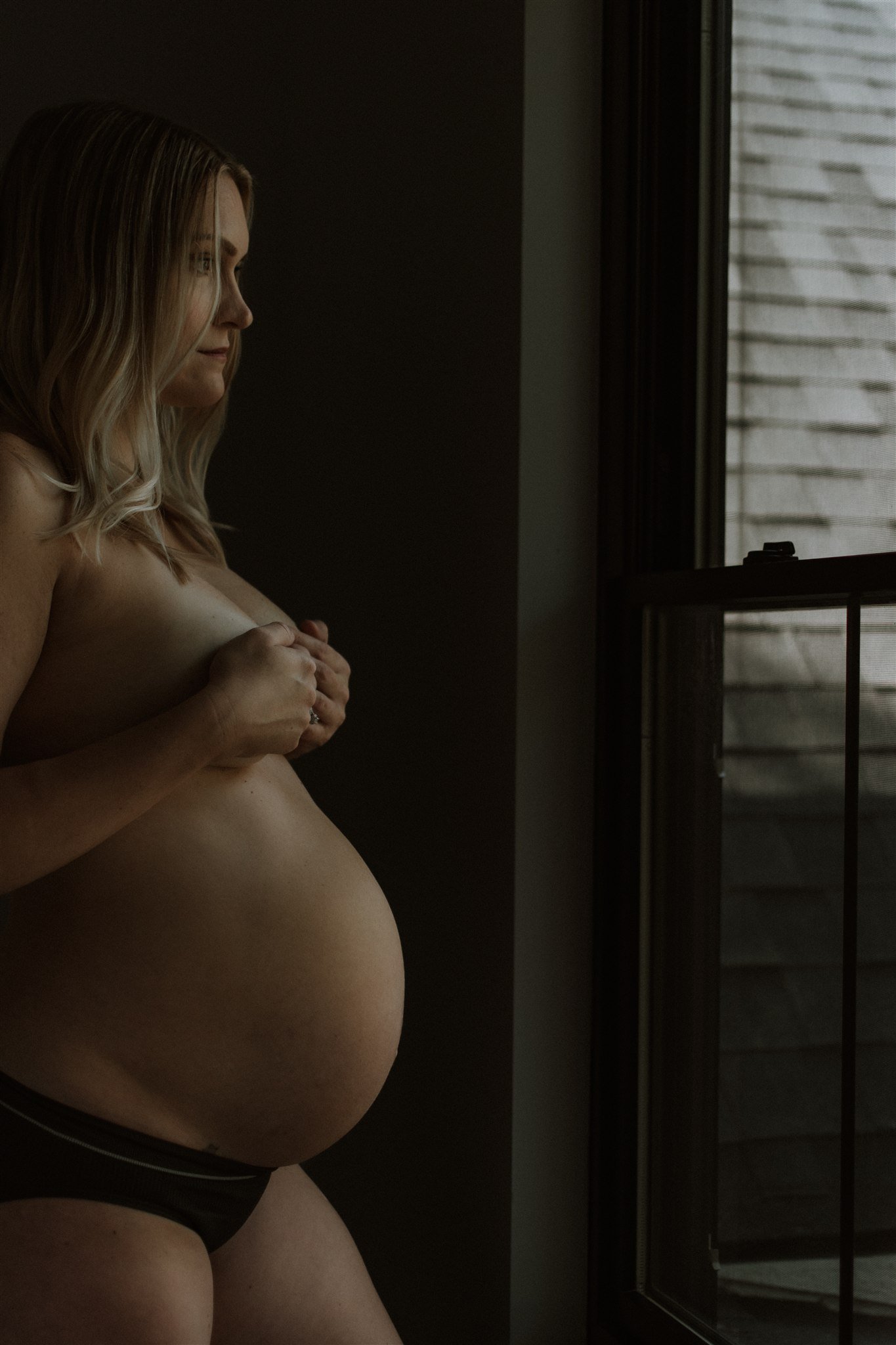 A pregnant woman standing by a window, looking outside with her hands holding her breasts.