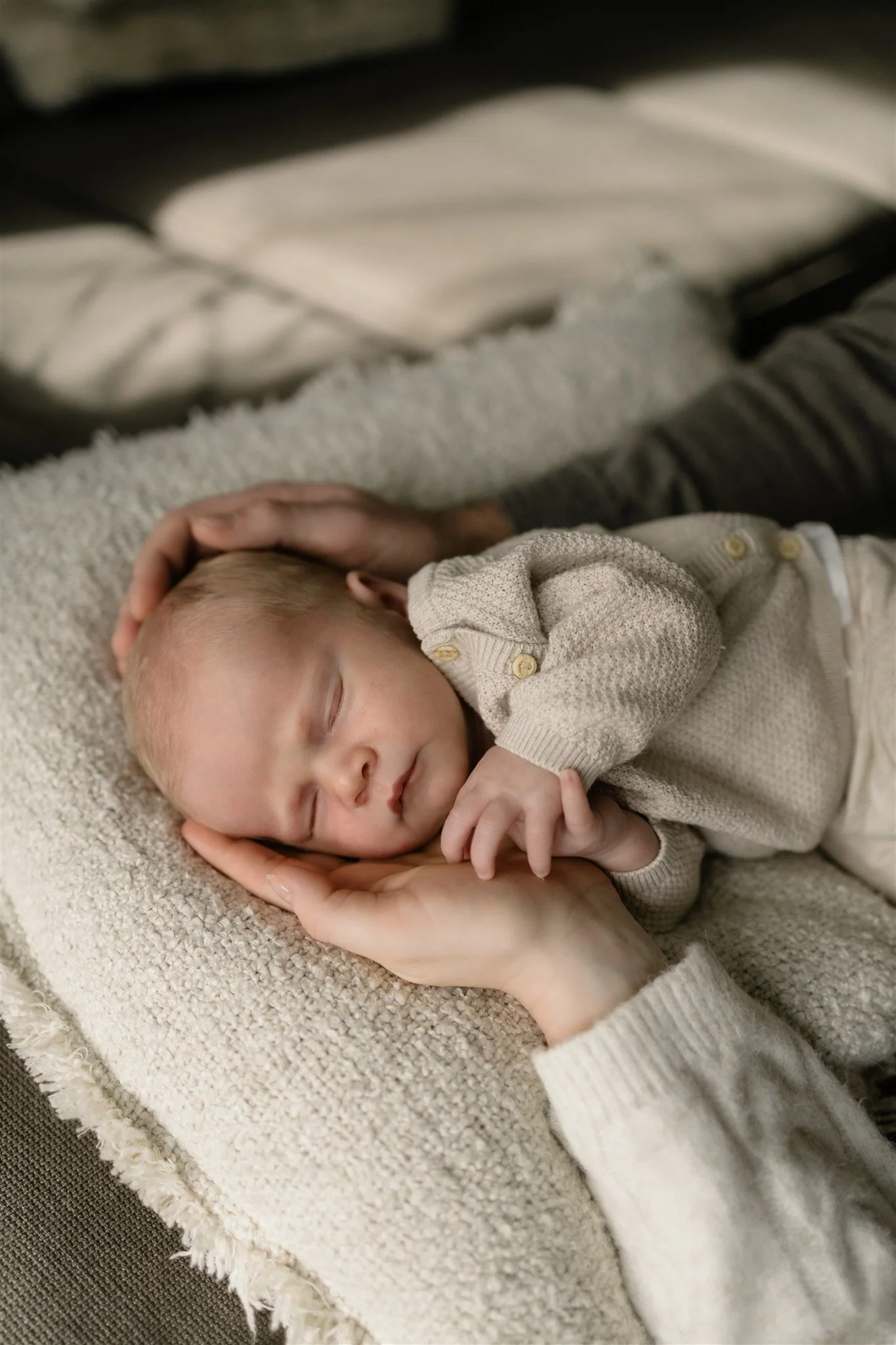 A newborn baby with blonde hair sleeping peacefully on a soft, textured blanket, gently held by an adult's hand.