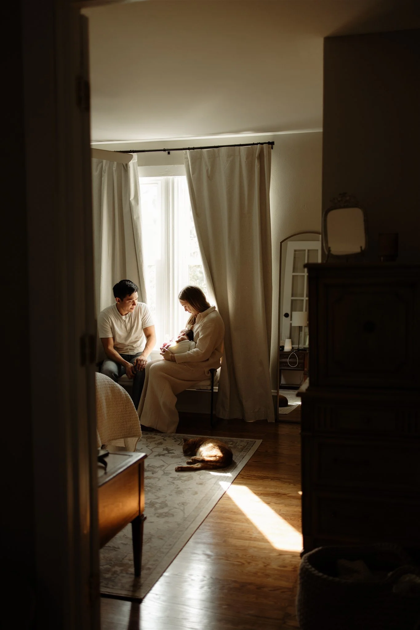 A couple sitting near a window with curtains, it looks like they are welcoming a newborn. There is a dog lying on a rug on the floor.