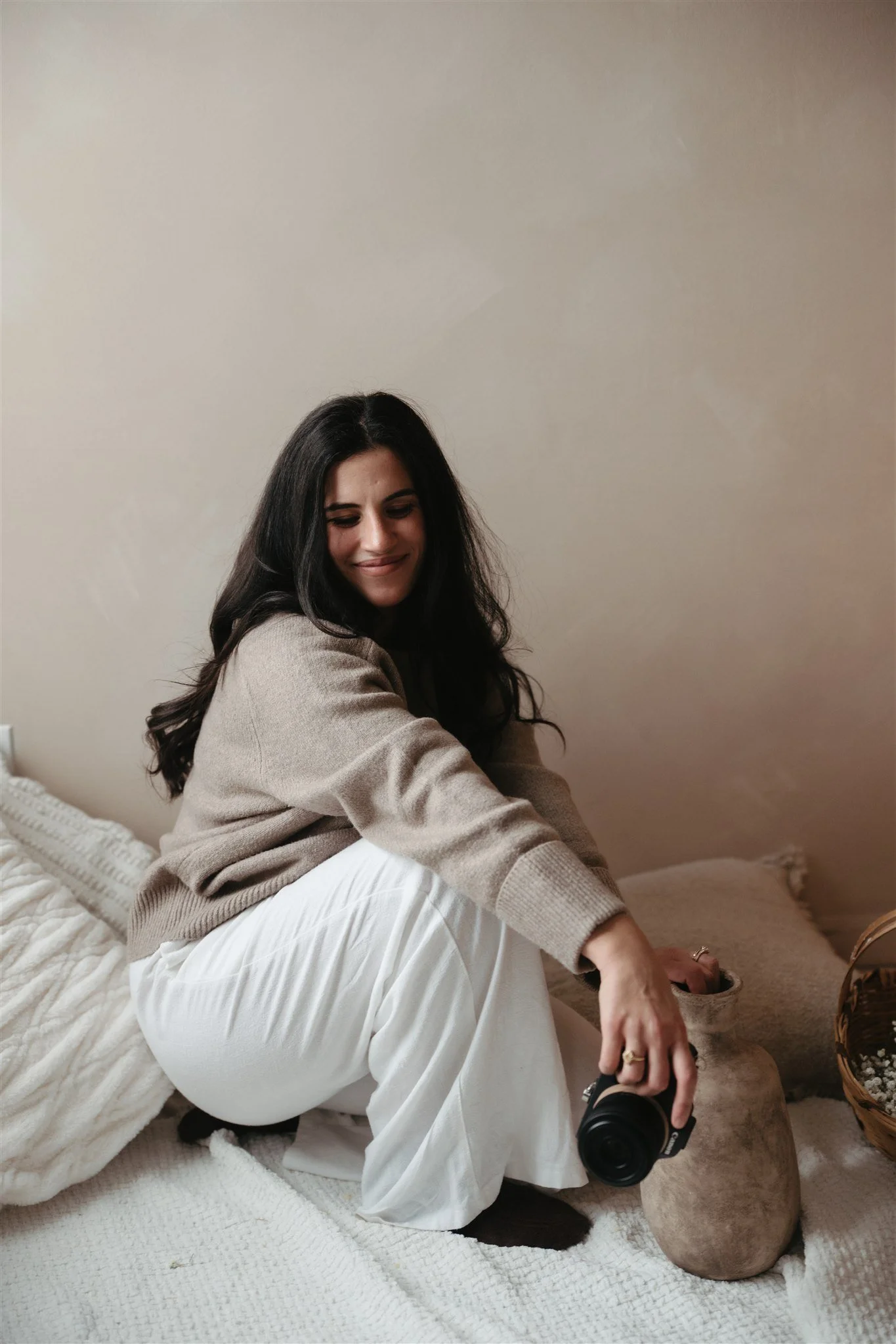 A woman with long dark hair smiling and sitting on a bed, holding a camera, in a cozy room with neutral-colored walls and soft bedding.