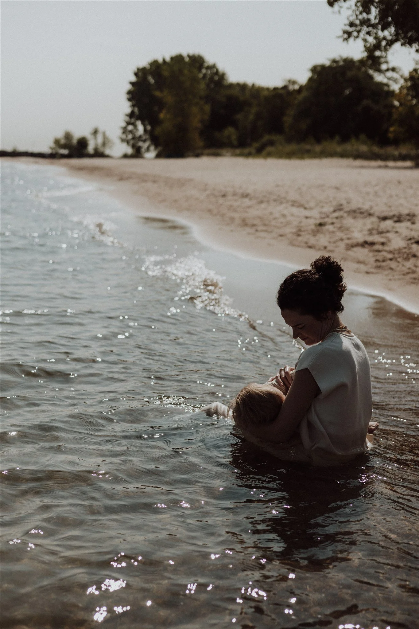 A woman holding a child in shallow water at the beach, with trees and sand in the background.
