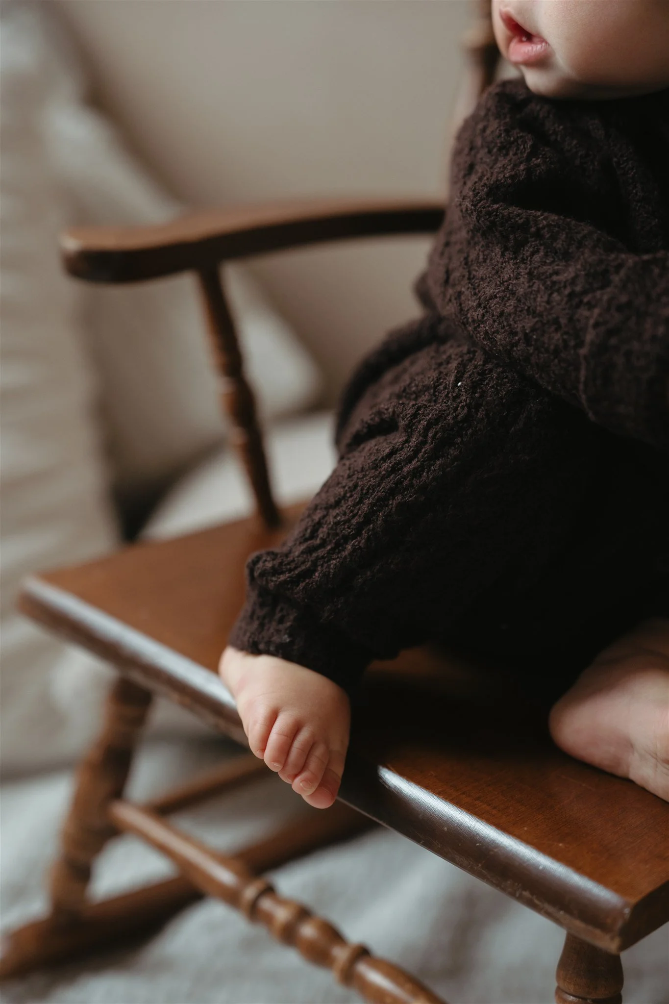 Close-up of a small child's hand resting on the edge of a wooden chair, with part of their face and mouth visible.