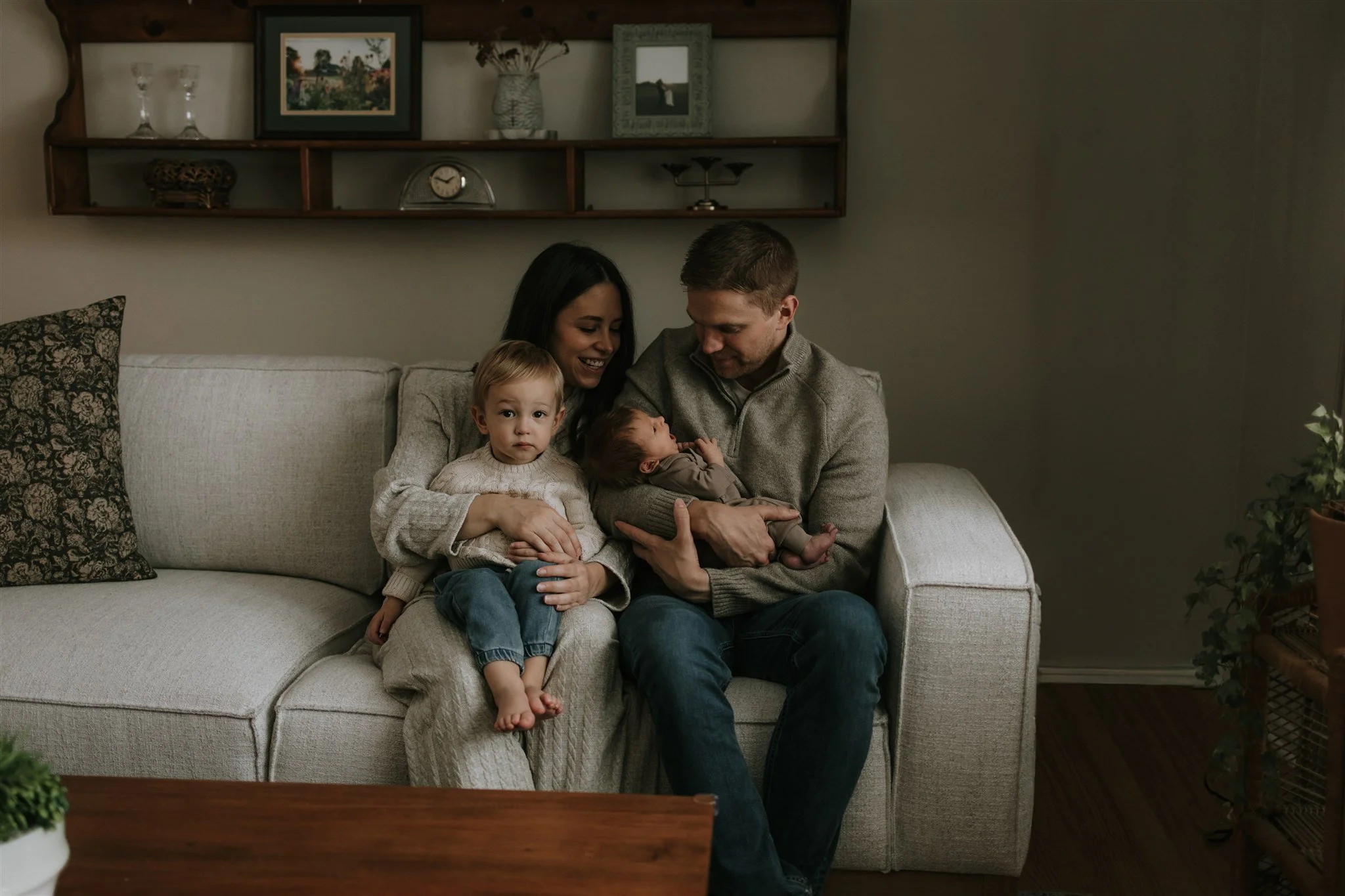 A family of four sitting on a beige sofa in a living room, with a woman, a man, a young girl, and a newborn baby, all smiling and looking happy.