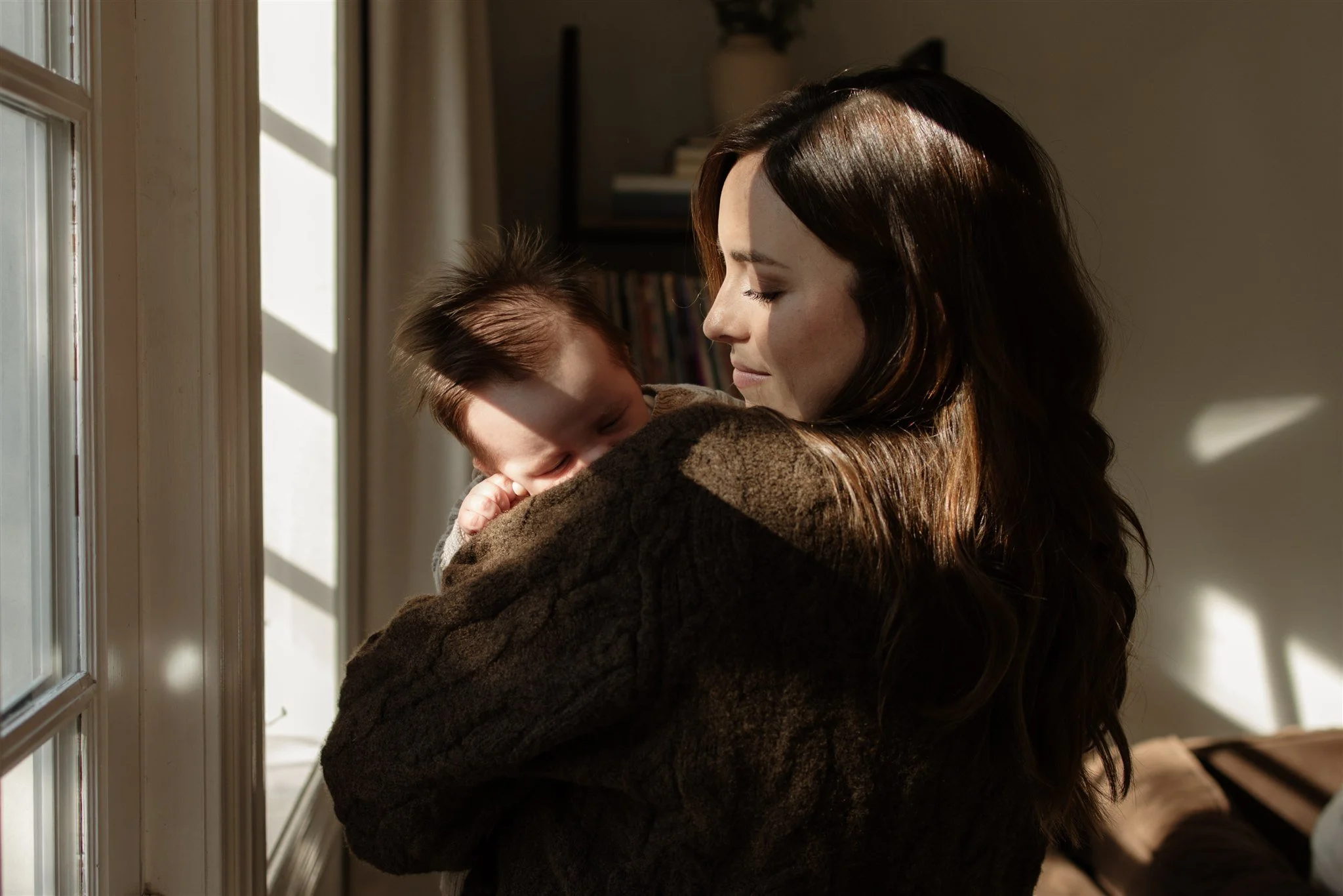 A woman holding a baby near a window with sunlight streaming in, creating shadows on the wall.