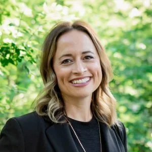 Professional headshot of Bekah Riker smiling outdoors in natural light, wearing a black blazer and dark top with greenery softly blurred in the background.