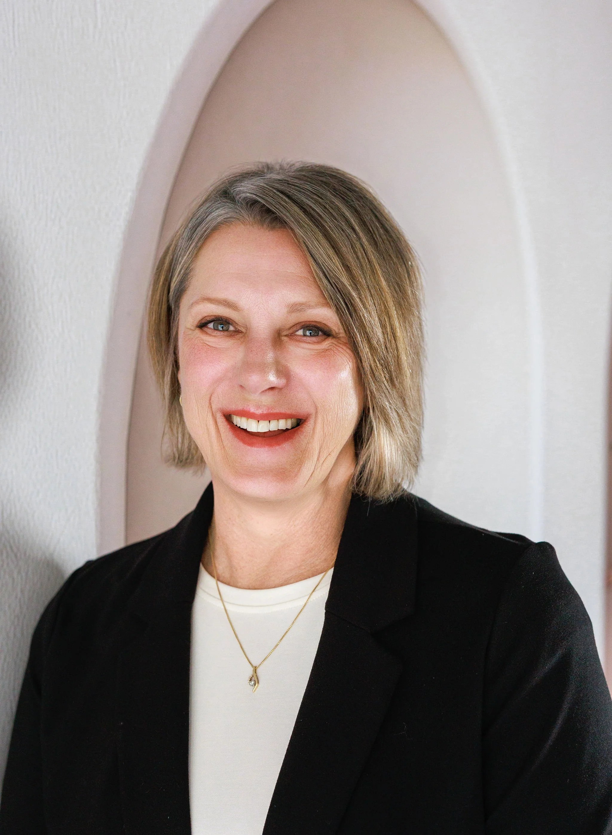 Close-up of a smiling woman with short brown hair, wearing a black blazer, white top, and a gold necklace, against a neutral background with a curved arch.