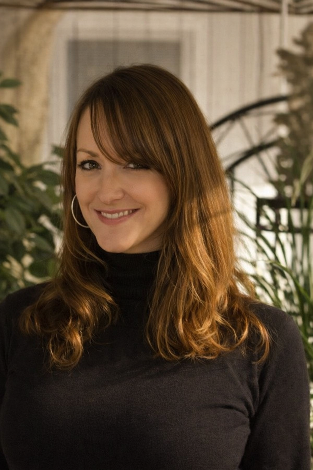 A woman with shoulder-length red hair smiling, wearing a black turtleneck sweater, standing in a room with plants and a window in the background.