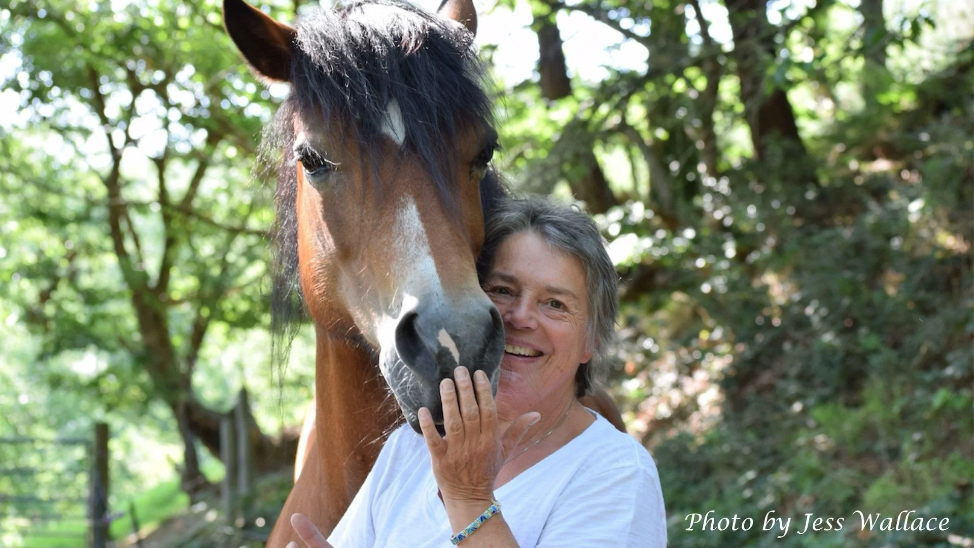 A woman with short gray hair smiling, holding hands on a horse's face, with a horse's head close to her face in a green outdoor setting.