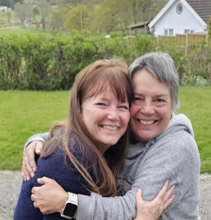 Two women hugging and smiling outdoors on a grassy lawn with trees and a white house in the background.