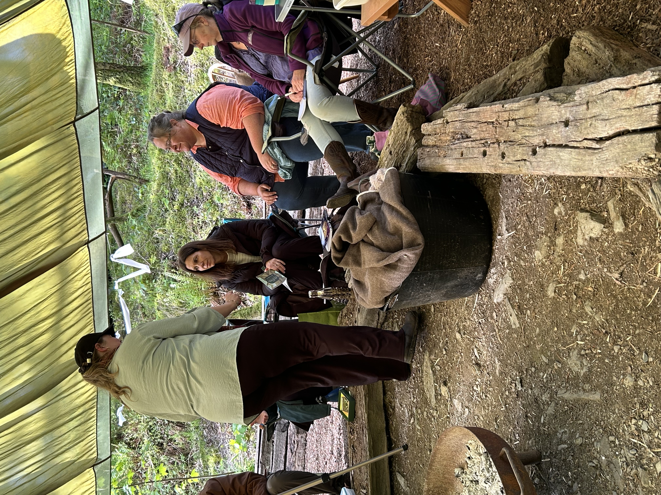 A group of five women sitting and standing around a campsite table, engaged in a discussion or activity, outdoors in a wooded area.