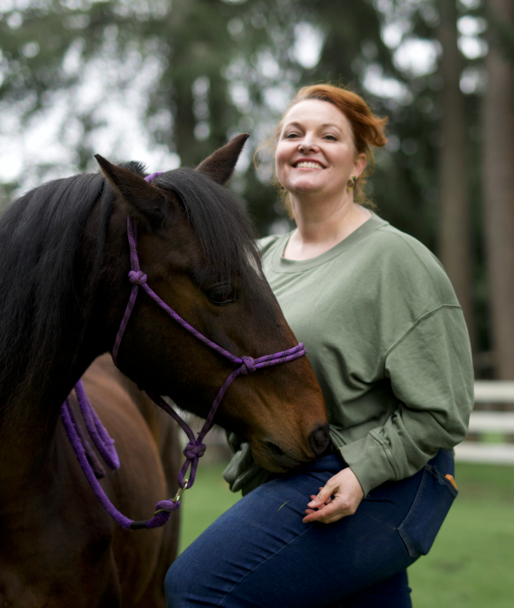 A woman smiling happily standing next to a brown horse with purple riding halter, outdoors with trees in the background.