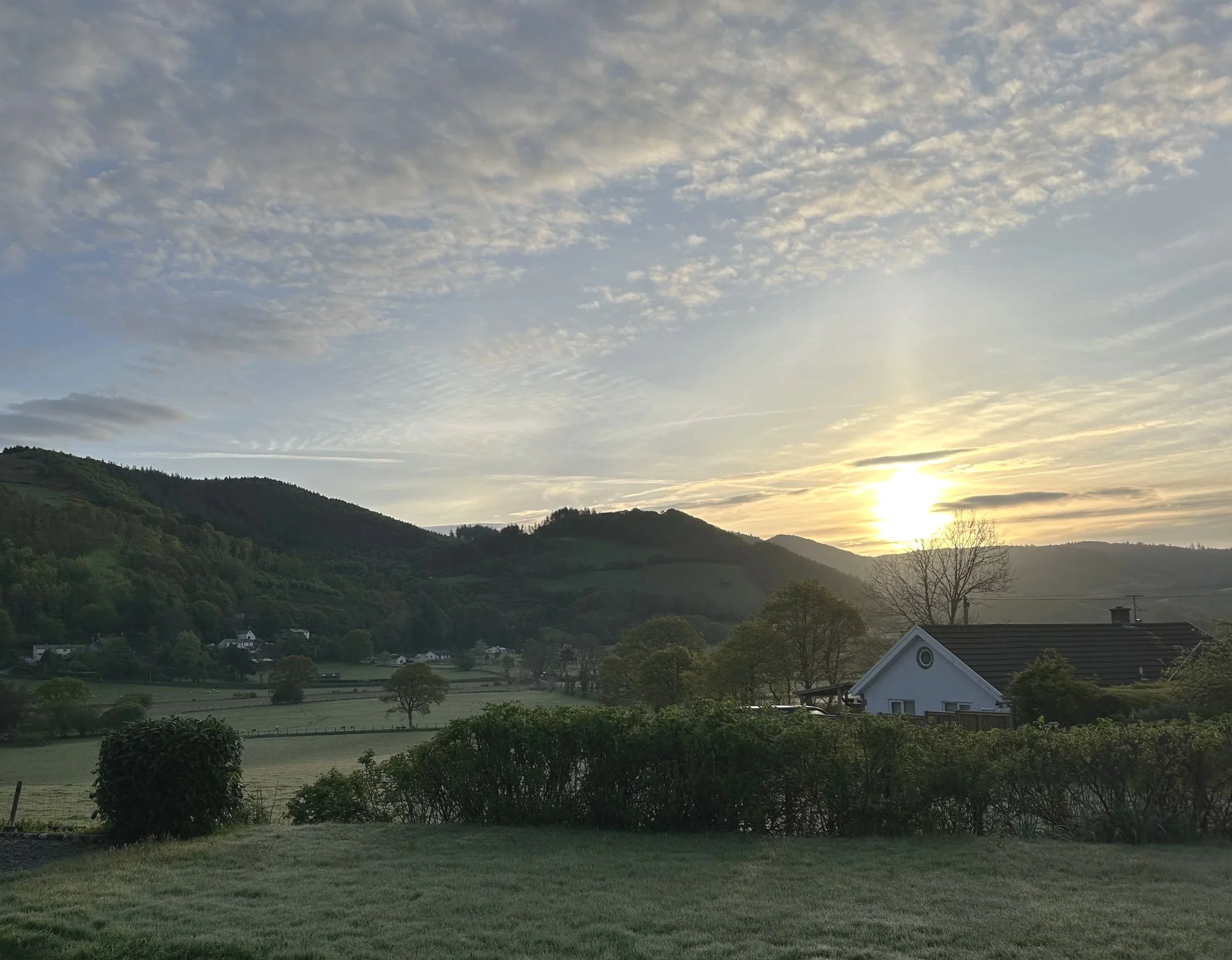 Scenic view of rolling green hills and a valley at sunset with a partly cloudy sky and a white house with a dark roof in the foreground.