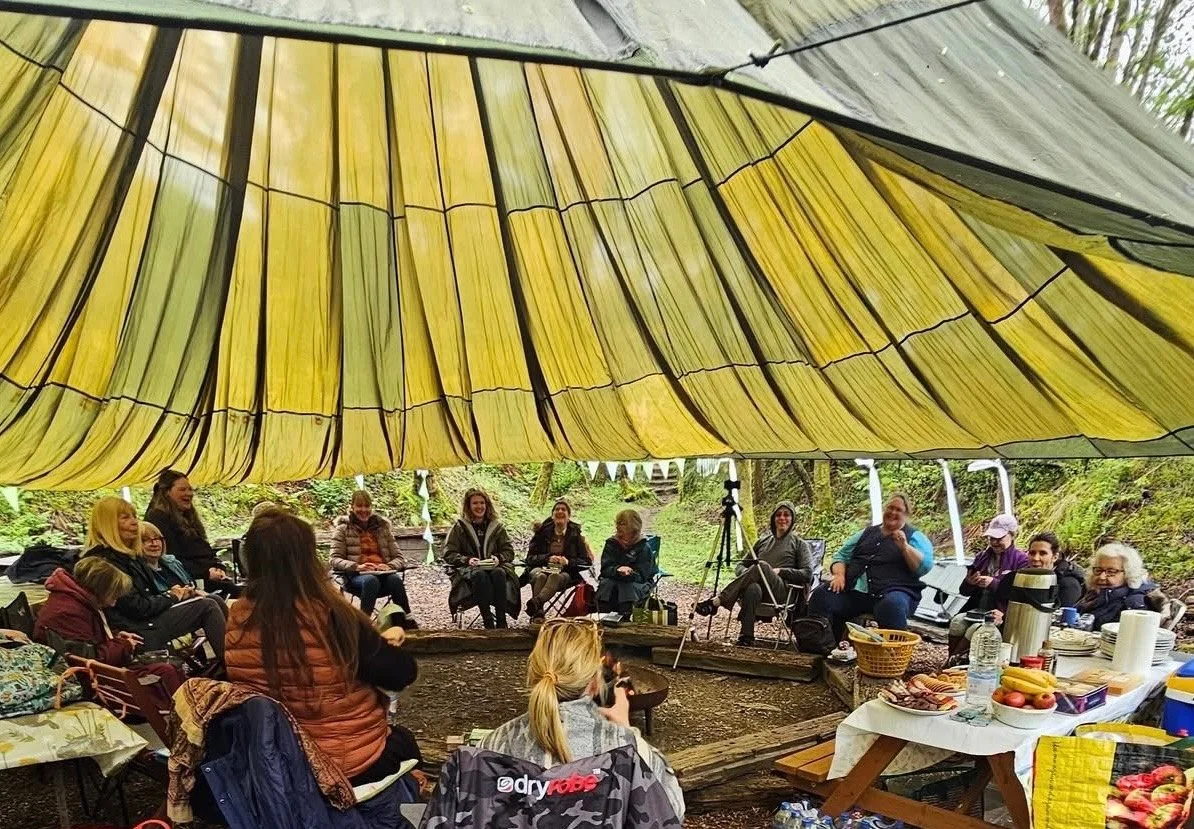 A group of people sitting in a circle under a large yellow and gray tent outdoors, with a table of food and drinks nearby.