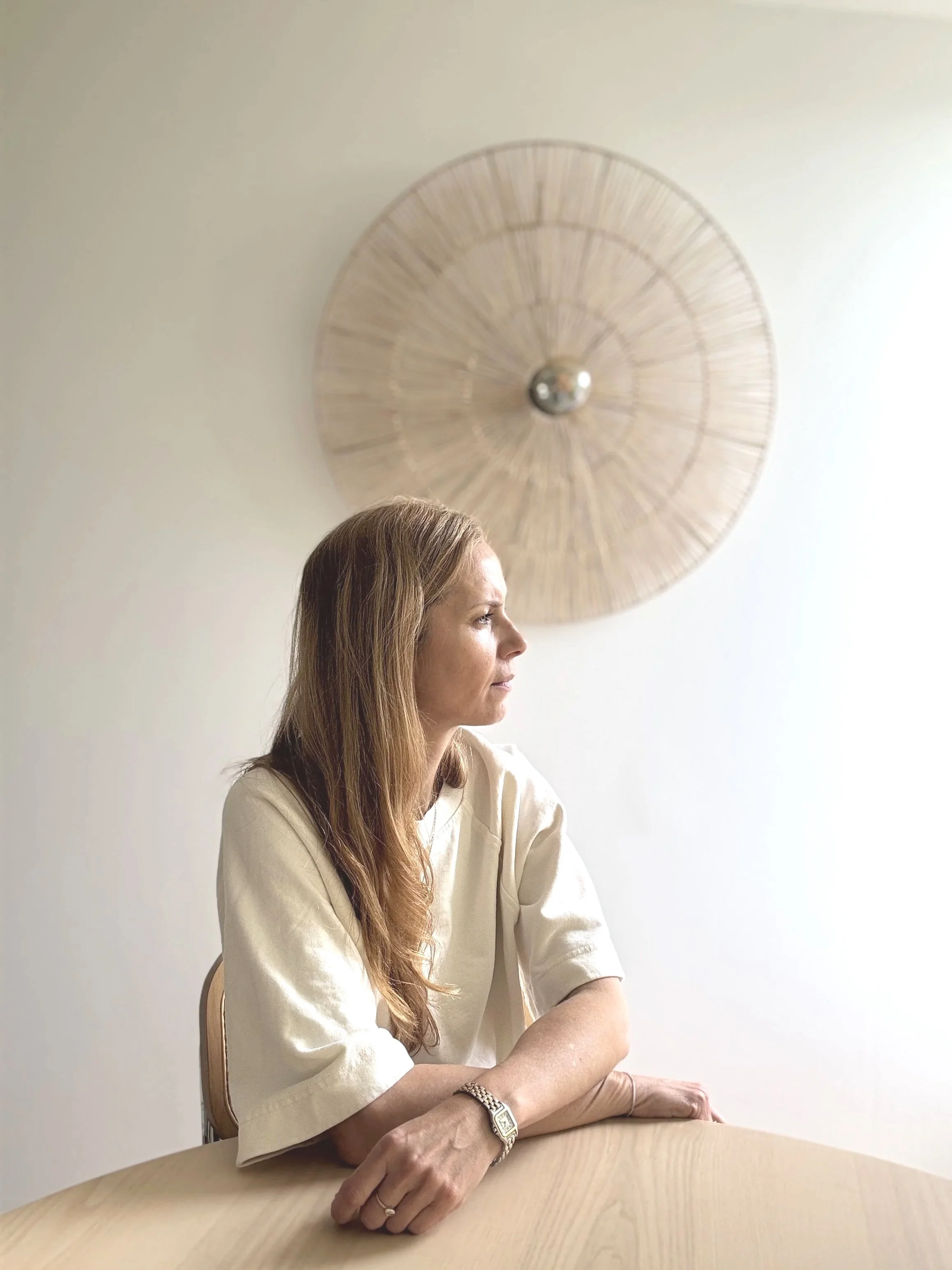 Une femme aux cheveux longs bruns, portant un chemisier beige, est assise à une table en bois clair. Elle regarde vers la droite, dans un intérieur lumineux. En arrière-plan, un mur blanc avec une décoration en bois rond.