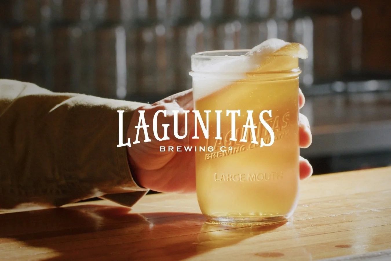 A person holding a glass of beer in a brewery setting. The beer has a frothy head and the glass has the Lagunitas Brewing Company logo on it.