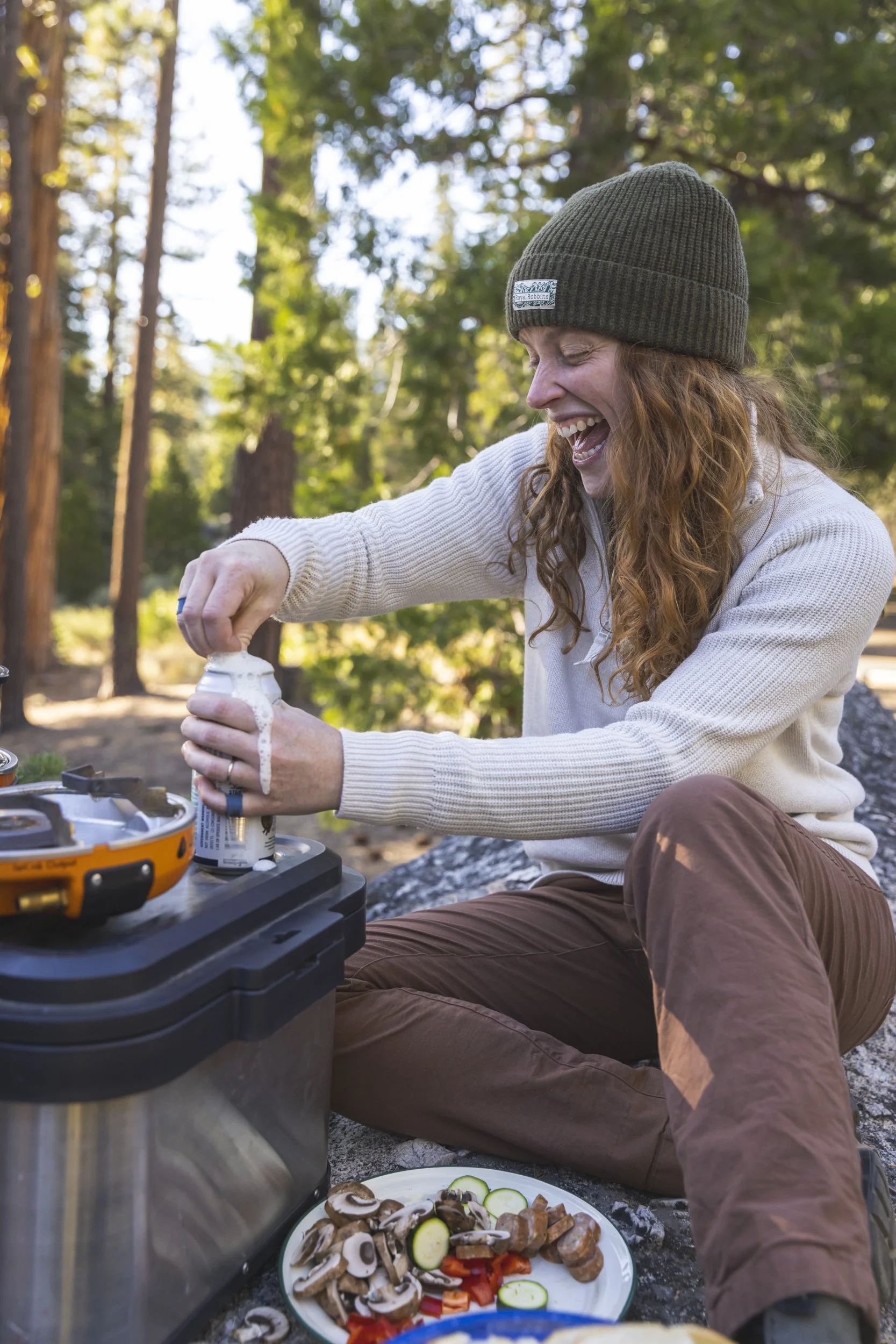 Woman with long red hair, wearing a knit hat and sweater, sitting outdoors in a forest and opening a can of food, part of a Dometic storage and cooking scene, photographed by Fieldborn Creative.