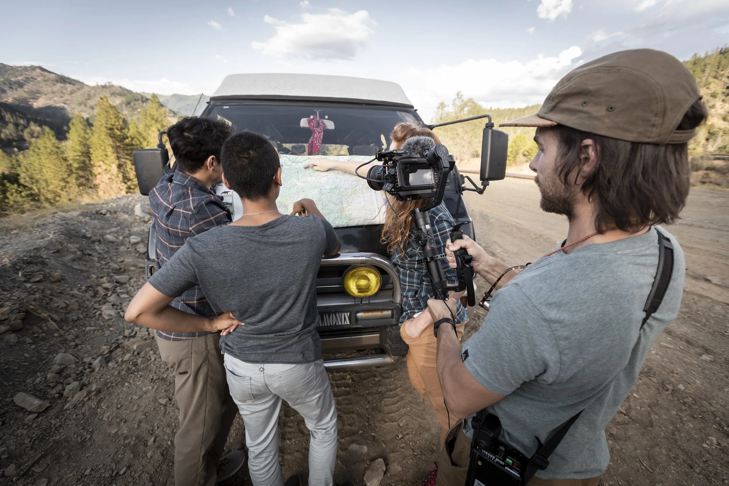 Behind-the-scenes photo of Kaare Iverson holding a camera in Humboldt County during an Osprey lifestyle shoot, with three people reviewing a map on the hood of a van, photographed by Fieldborn Creative.