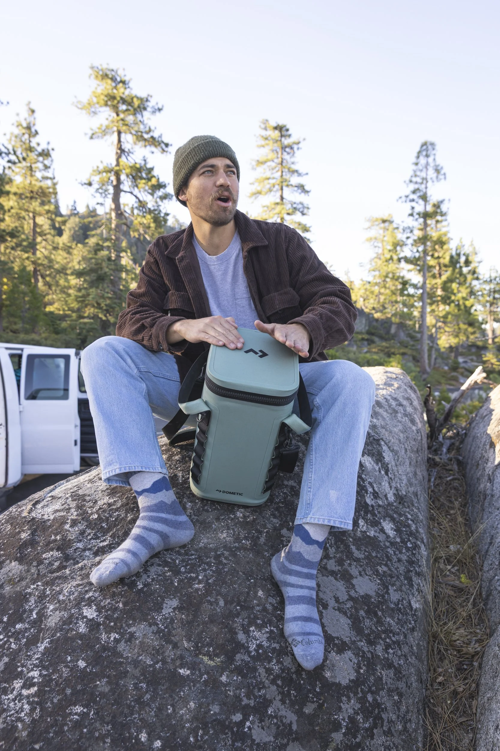 Man sitting on a large rock in a forest, wearing a beanie, brown jacket, light blue jeans, and gray striped socks, using a light green insulated Dometic cooler as a drum while singing, with trees and a clear sky in the background, photographed by Fie