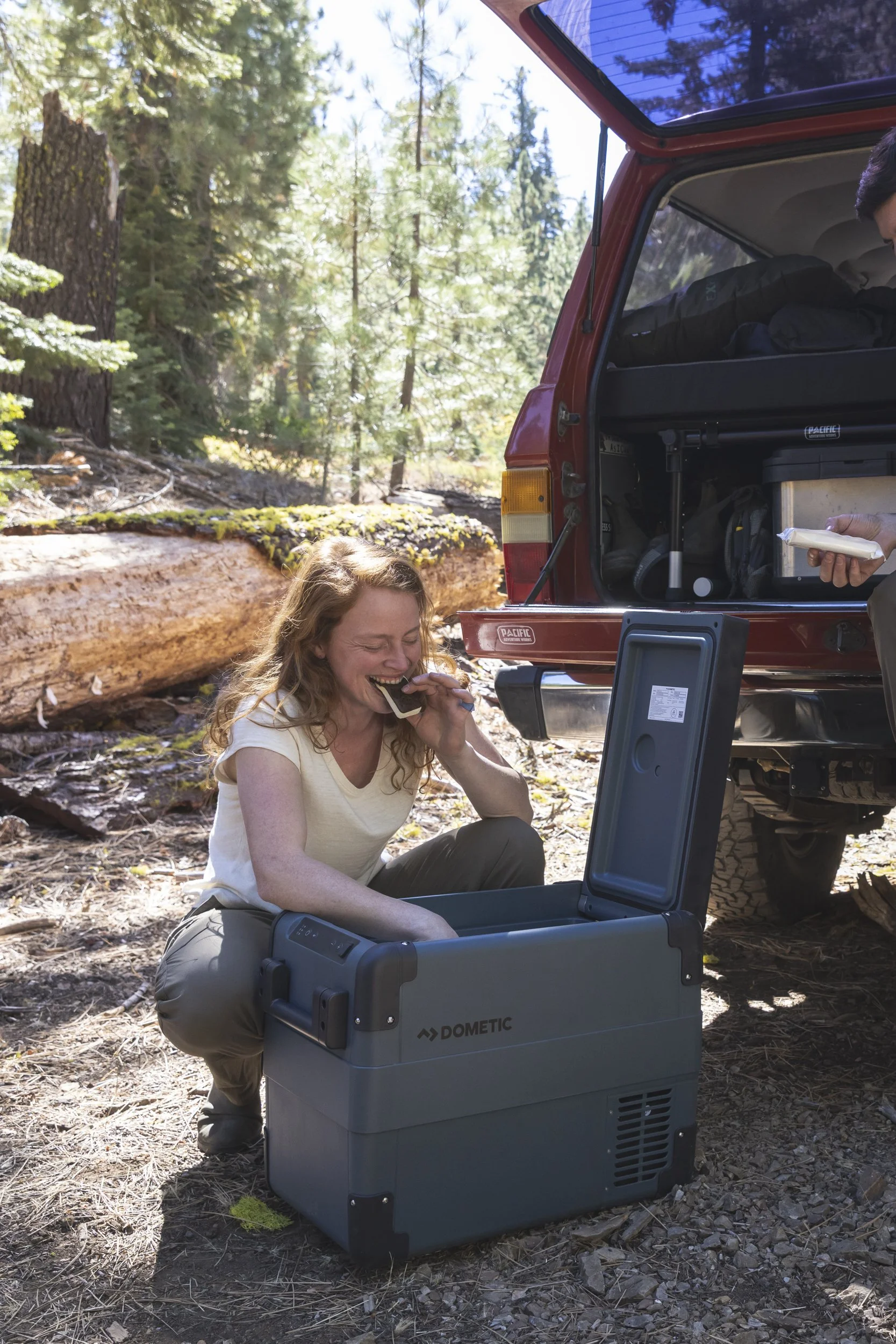 Woman sitting on the forest ground in front of an open portable Dometic cooler, laughing and eating an ice cream sandwich, with trees in the background and the open trunk of a red SUV nearby, photographed by Fieldborn Creative.