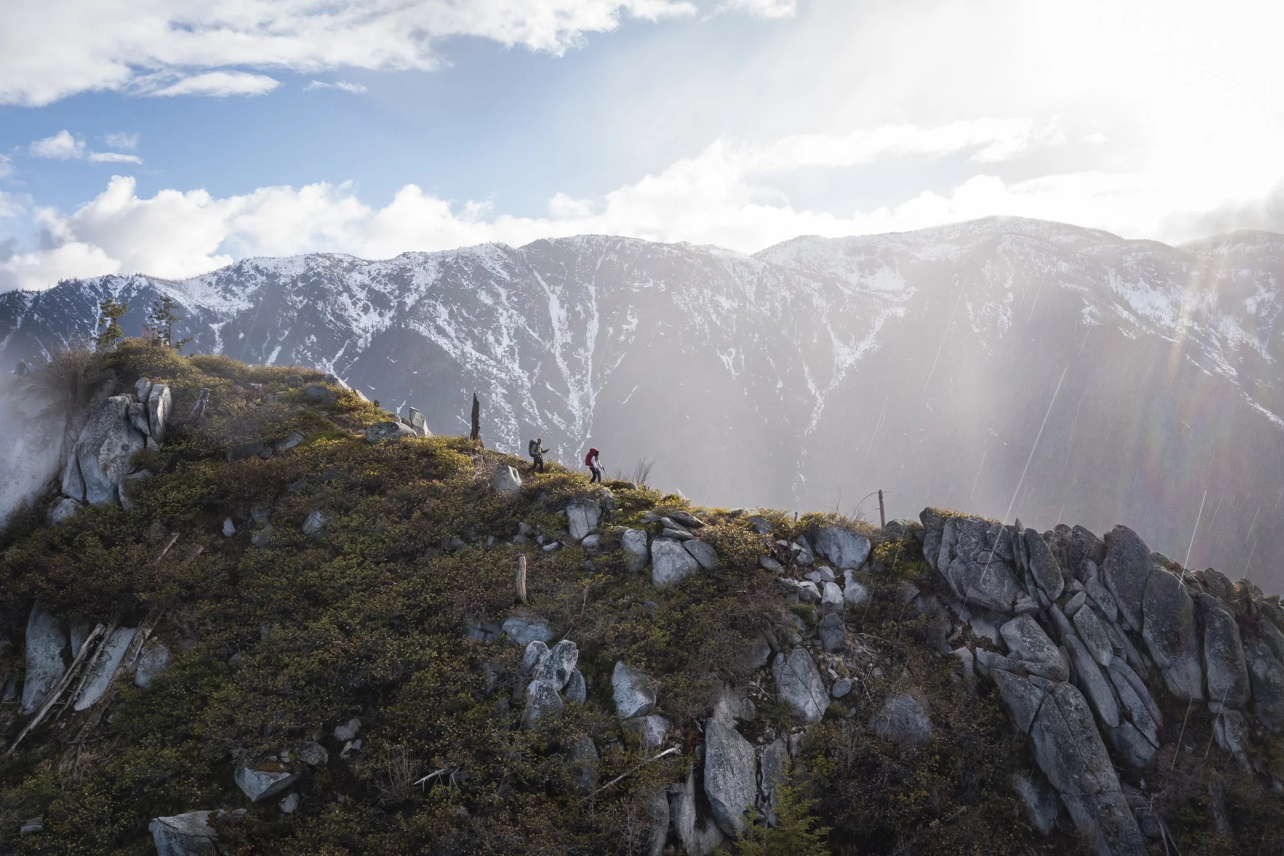 Hikers trekking up a rocky mountain ridge with snow-capped peaks in the background, under a partly cloudy sky with sunlight.