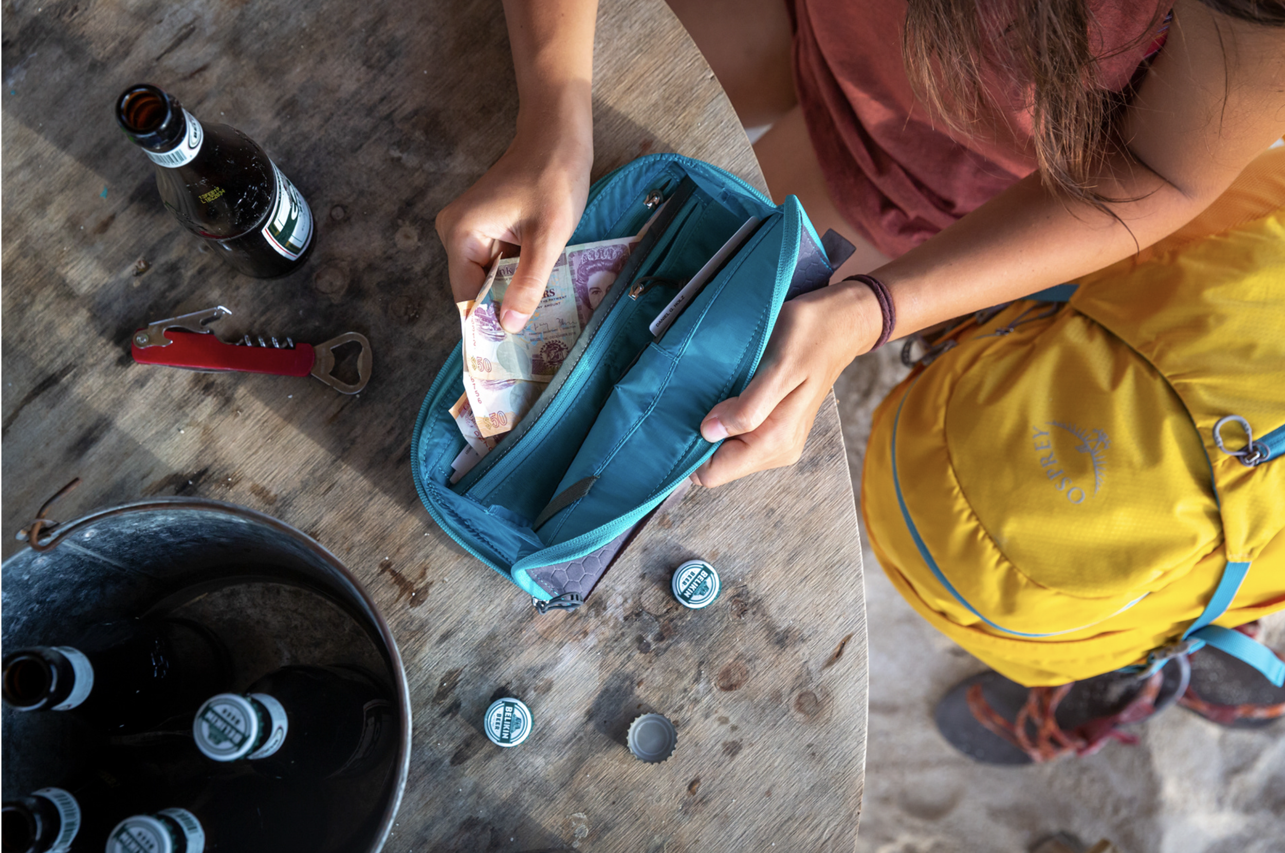 A person packing money into a turquoise wallet on a wooden table, with empty beer bottles, bottle caps, and a bottle opener nearby. The person is wearing a pink sleeveless top and yellow backpack, with only part of their arm and body visible.  