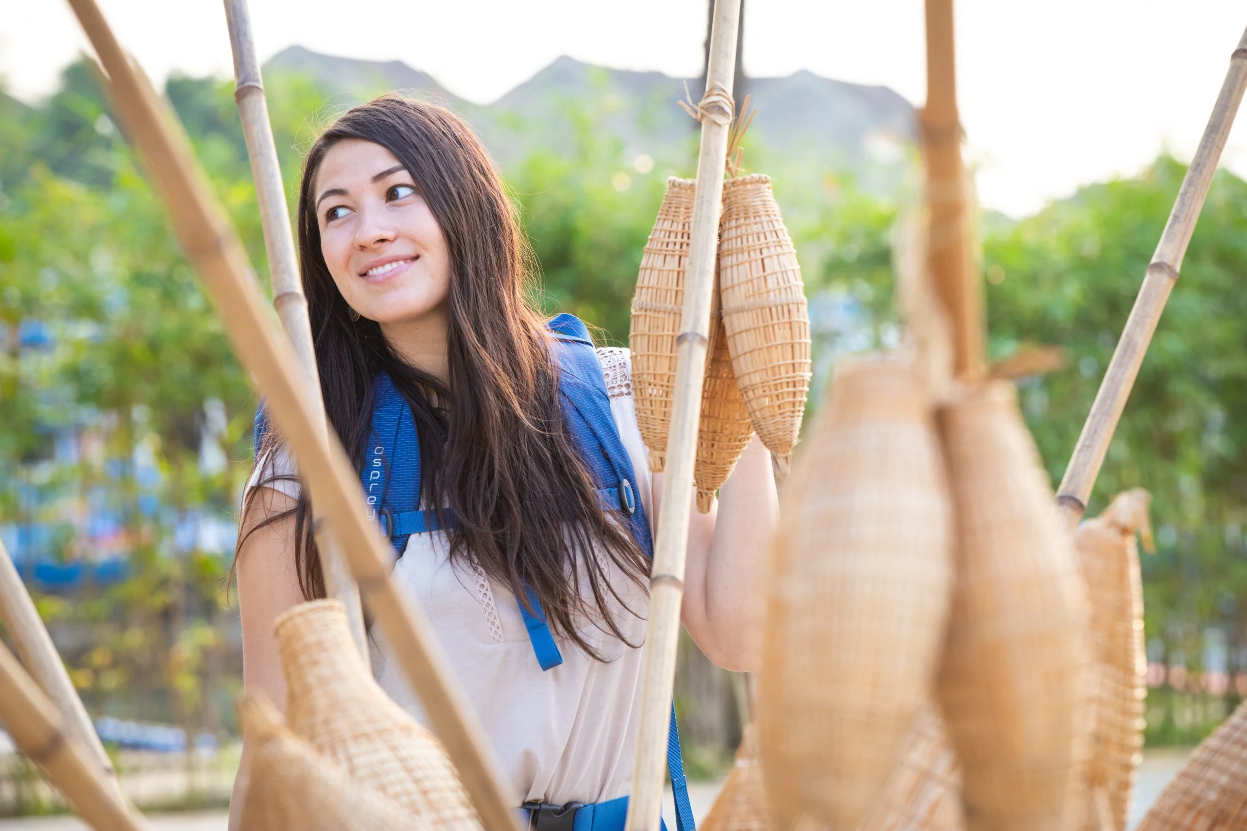 A young woman with long dark hair and a blue backpack standing among bamboo structures outdoors, smiling softly with a blurry green background and mountain in the distance. Commercial lifestyle production by Fieldborn Creative.